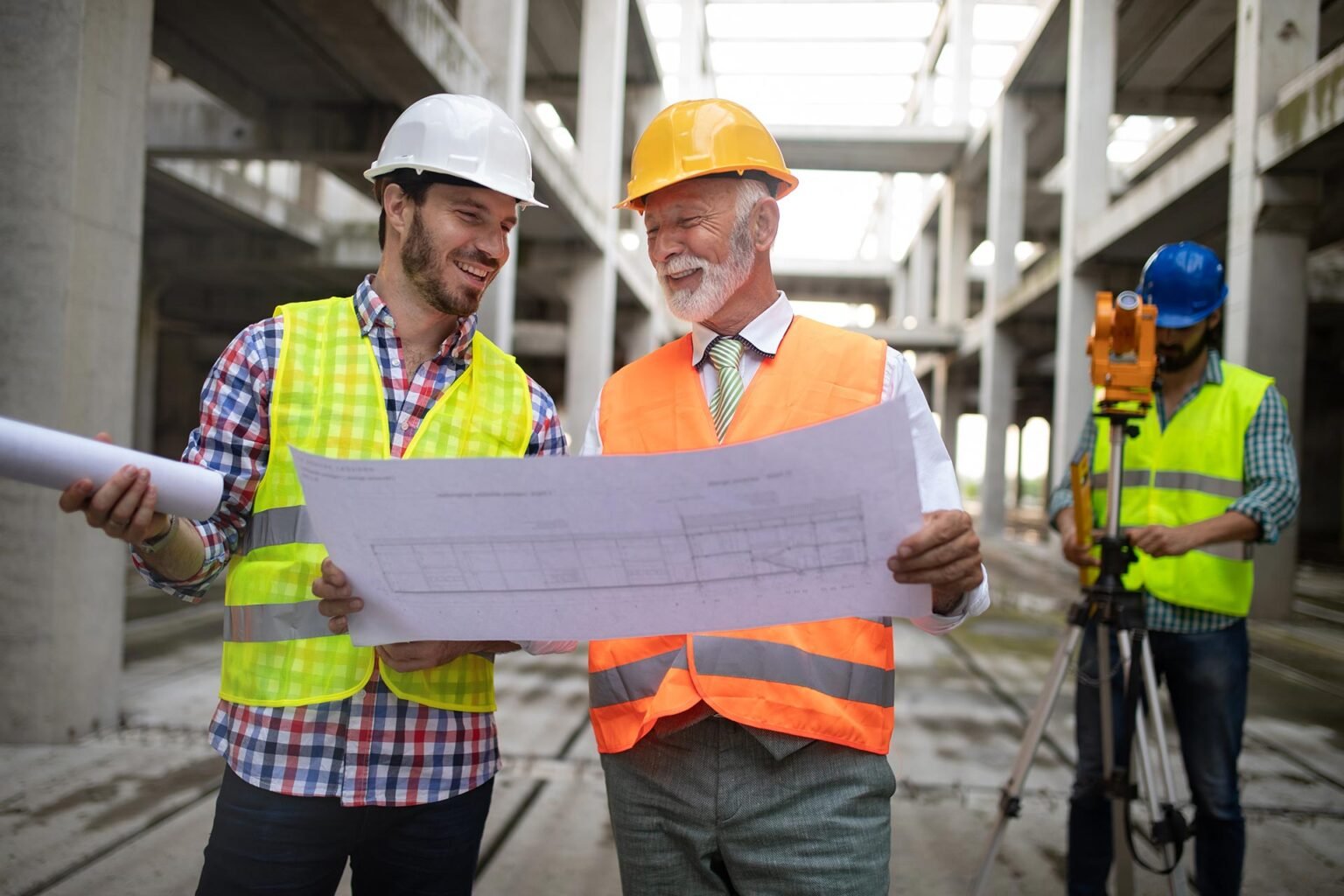 An engineer and a foreman reviewing a drawing on site while a surveyor works a total station behind them.